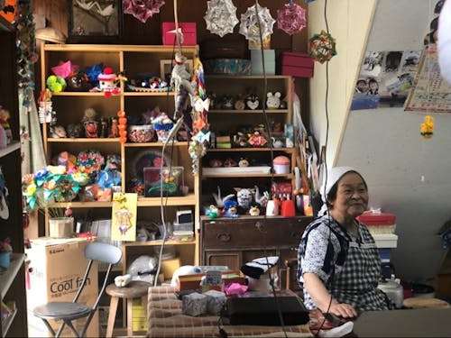 折り紙が並ぶ店内と座る信さん